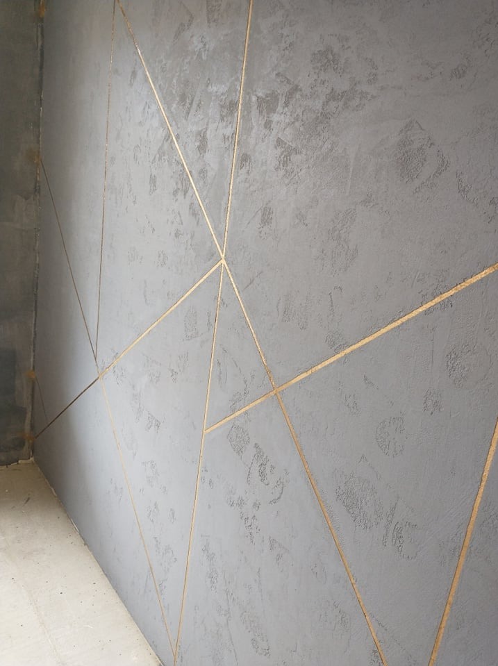 Szara ściana z dekoracyjną fakturą i złotymi liniami tworzącymi geometryczny wzór, widok z boku, surowy beton w tle.