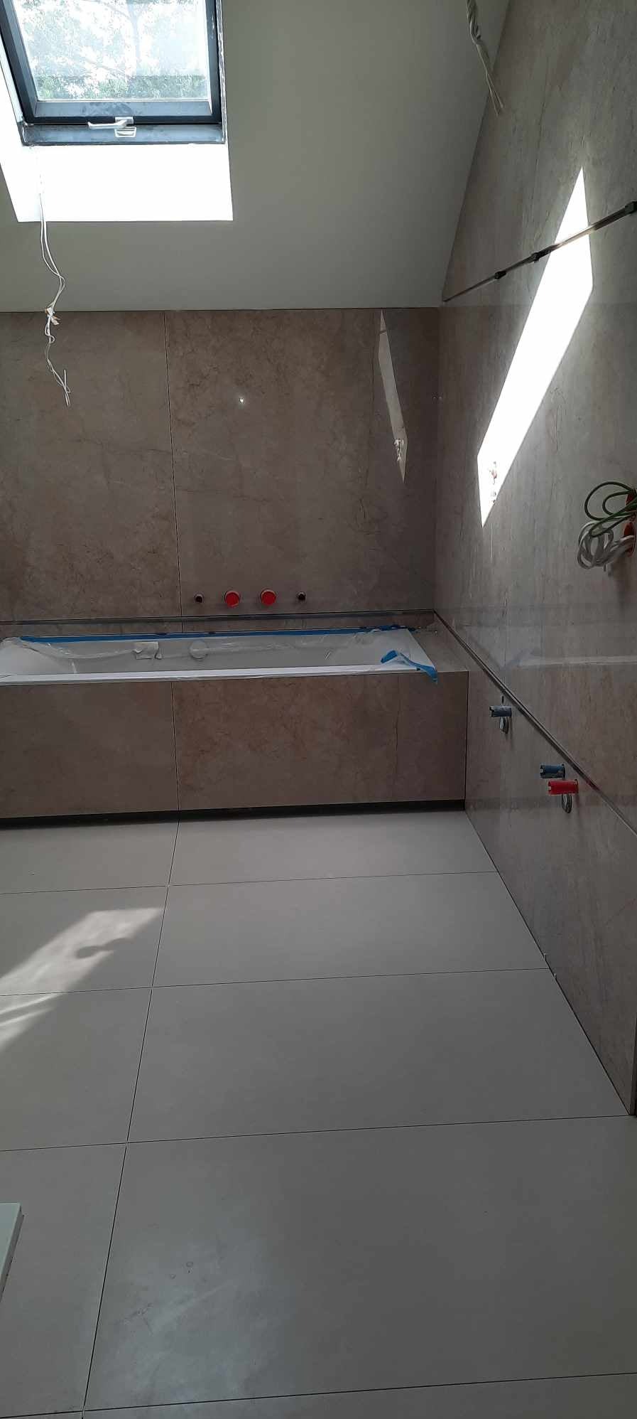 Wykończona łazienka z beżowymi płytkami, białą wanną zabezpieczoną folią i oknem dachowym. Widoczne przyłącza wodne i fragment instalacji elektrycznej.