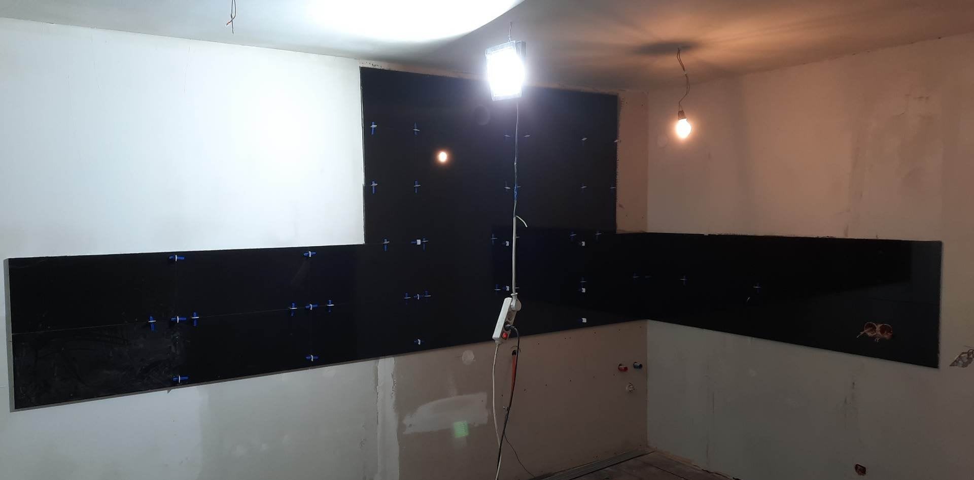 Czarne, błyszczące płytki ścienne w trakcie montażu w kuchni, z krzyżykami dystansowymi, oświetlone lampą budowlaną. Widoczne surowe ściany i instalacje.