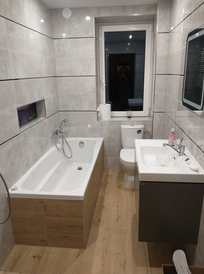 Jasna łazienka po remoncie: wanna obudowana drewnopodobnymi płytkami, umywalka z szafką, toaleta, lustro i okno. Płytki na ścianach w odcieniach szarości z czarnymi fugami.