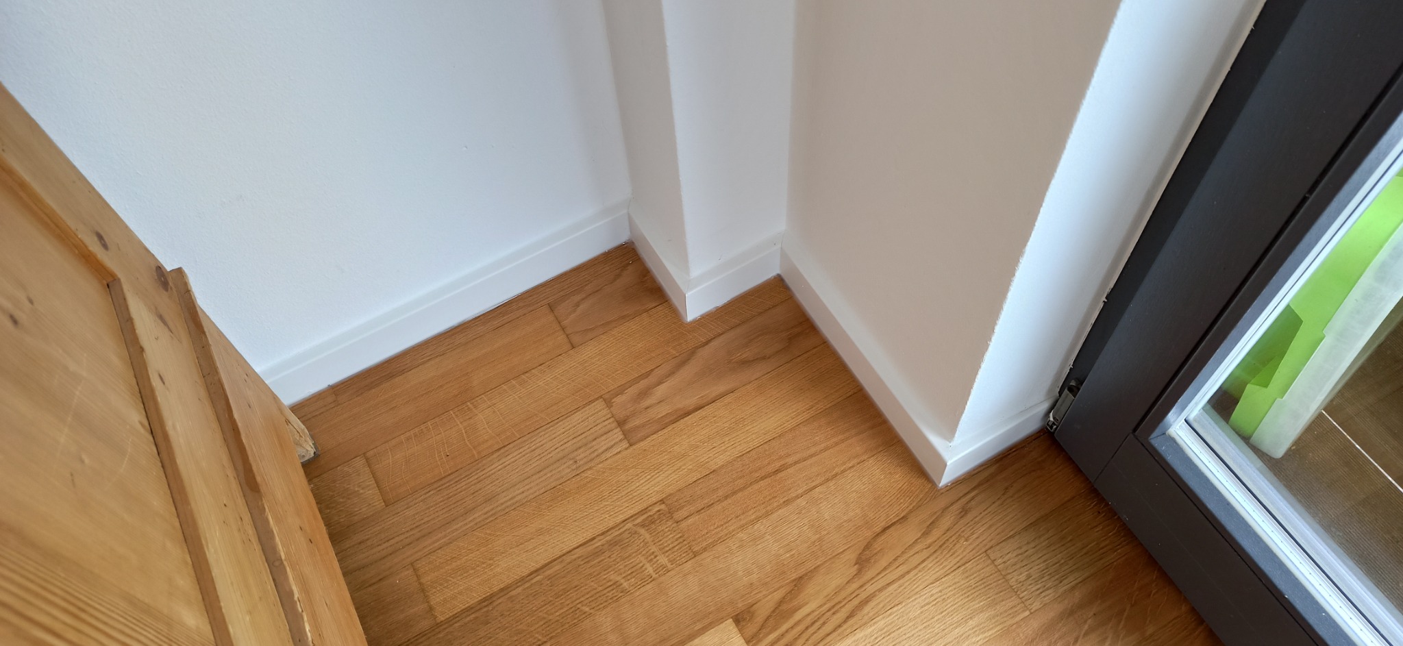 Parkiet dębowy ułożony w jodełkę, białe listwy przypodłogowe przy ścianie i drzwiach. Widok z góry, naturalne oświetlenie, detale wykończenia.