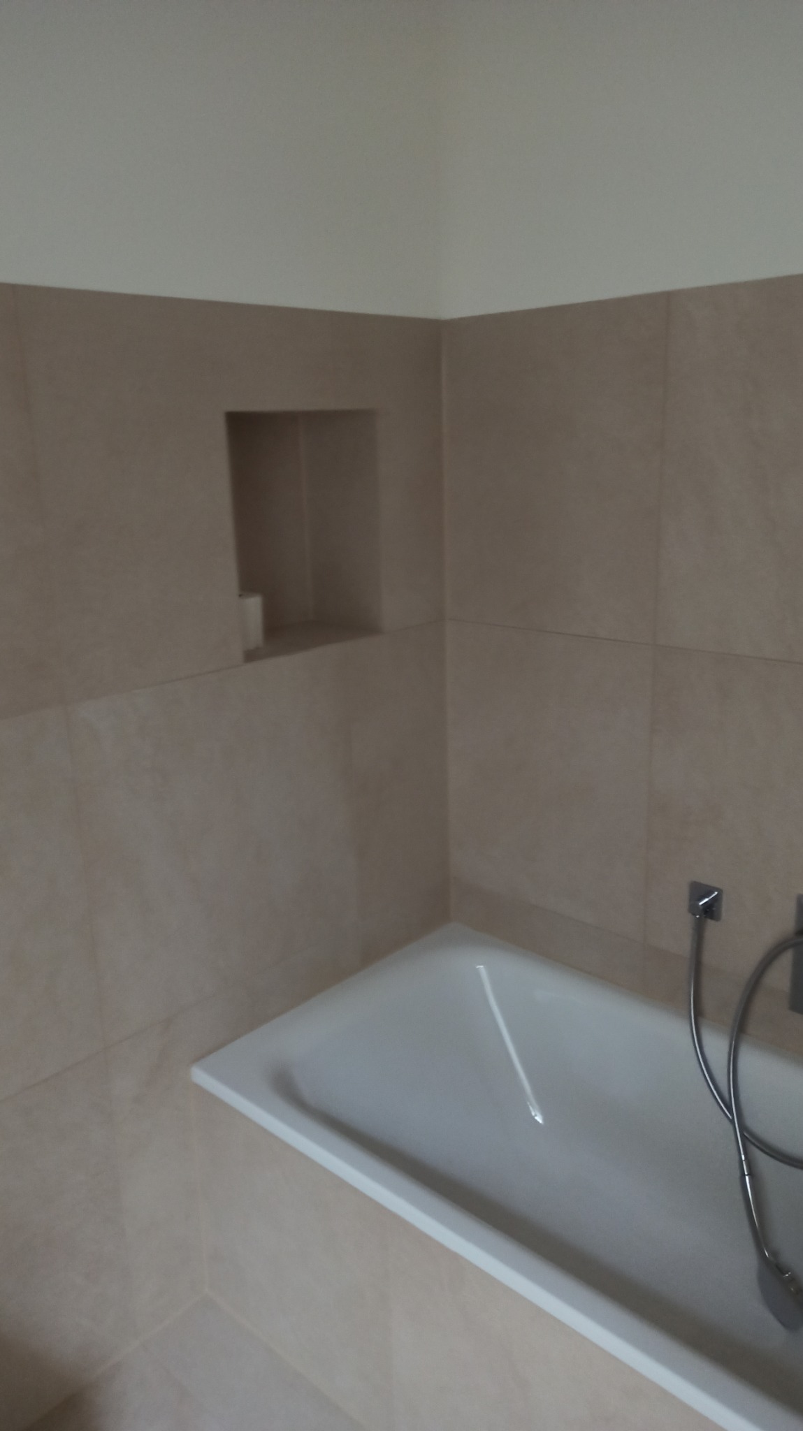 Beżowa łazienka po remoncie: wanna, płytki i wnęka w ścianie. Minimalistyczny design, jasne wnętrze. Widok na fragment baterii wannowej.