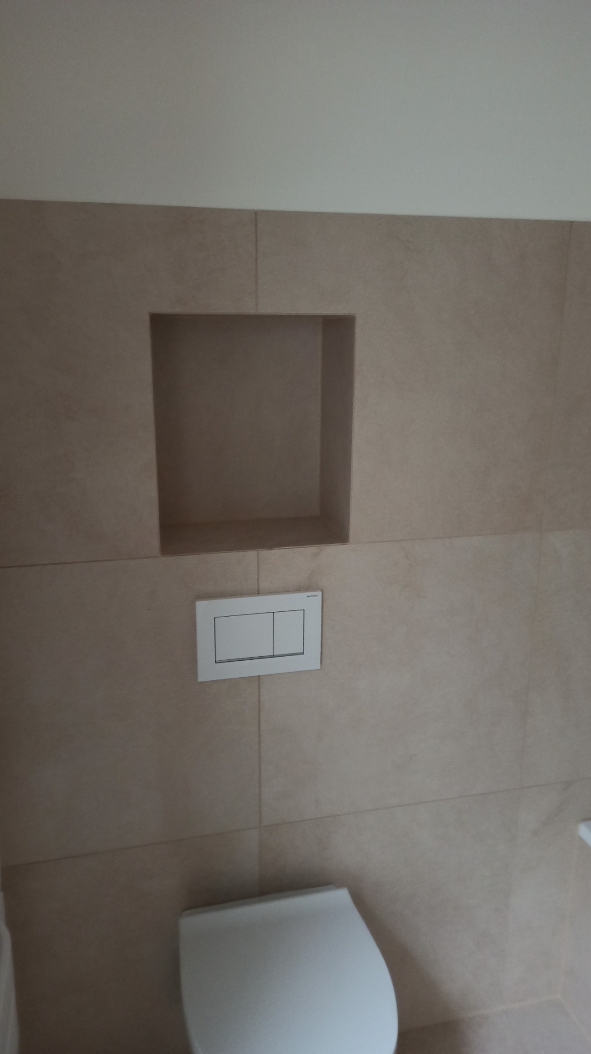 Beżowa, wykafelkowana ściana w łazience z wnęką i przyciskiem spłukującym nad toaletą. Minimalistyczny design, neutralna kolorystyka, nowoczesne wykończenie.