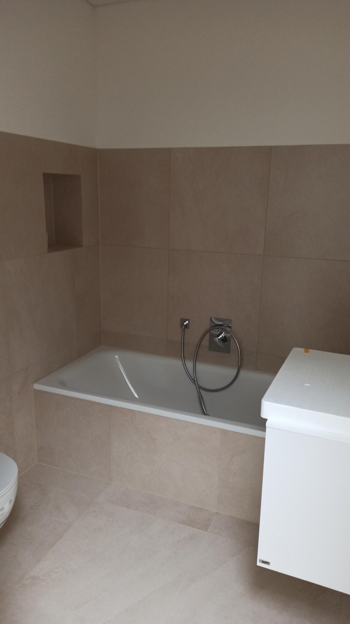 Beżowa łazienka po remoncie: wanna z baterią, zabudowa umywalki i płytki w ciepłym odcieniu. Widoczny fragment toalety. Minimalistyczny design.