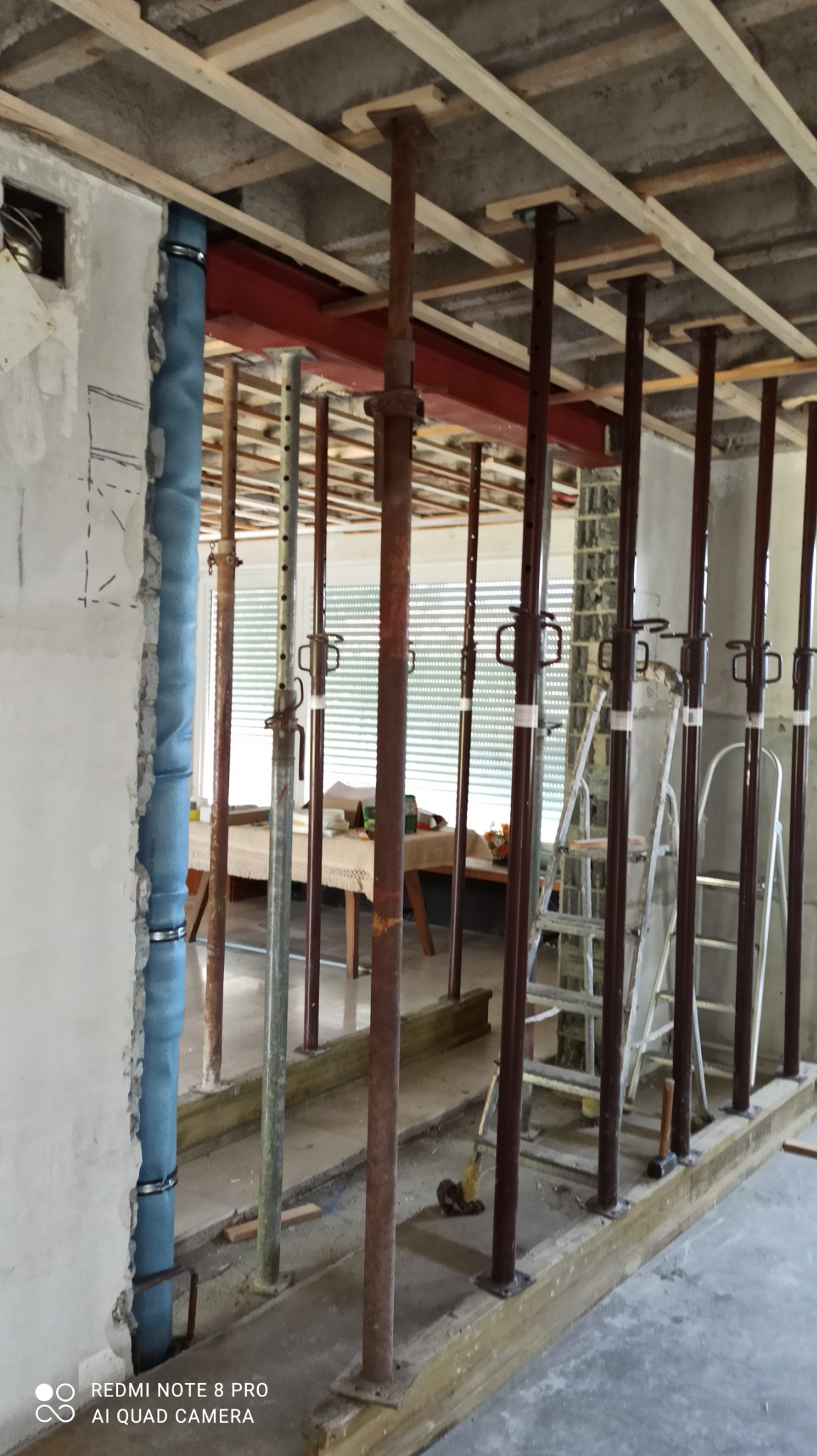 Wnętrze w trakcie remontu: podpory stropowe, instalacja w ścianie, surowy beton. Widoczne narzędzia i drabina. Prace przygotowawcze do wykończenia.