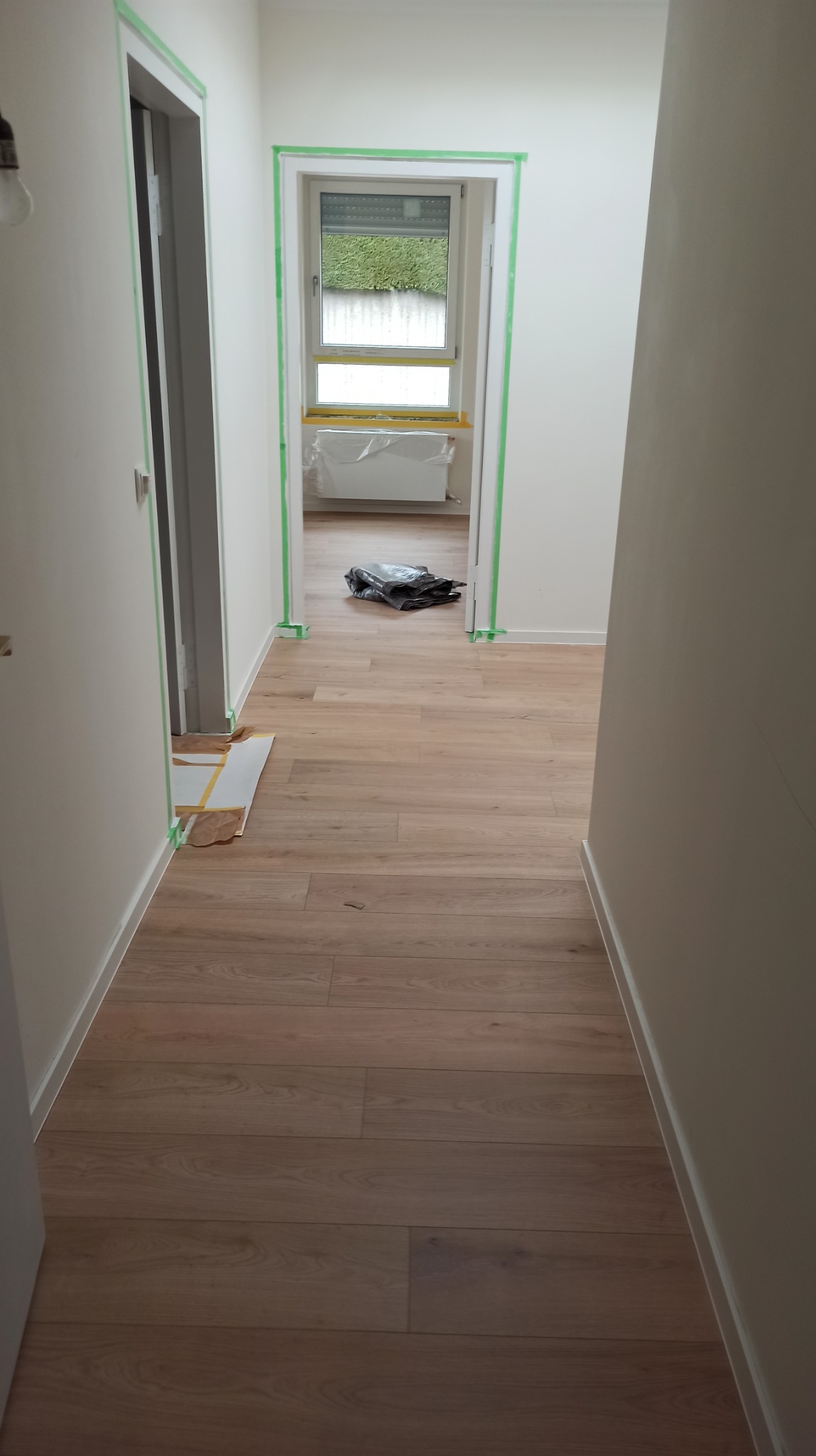 Jasny korytarz po remoncie z nową podłogą i świeżo pomalowanymi ścianami, zabezpieczonymi taśmą malarską. Widok na okno i grzejnik w tle.