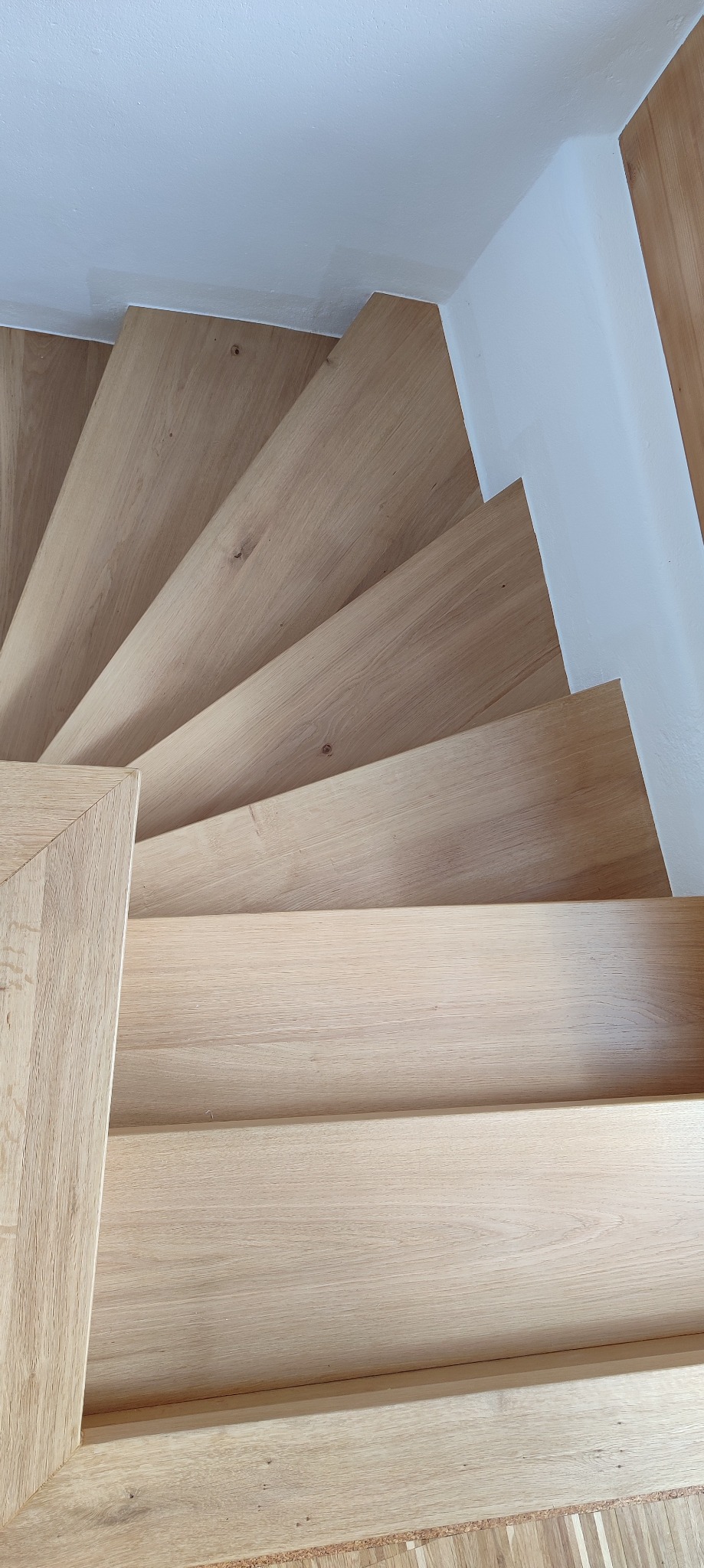 Jasne, drewniane schody spiralne z białymi ścianami, widziane z góry. Naturalne oświetlenie podkreśla teksturę drewna i minimalistyczny design.