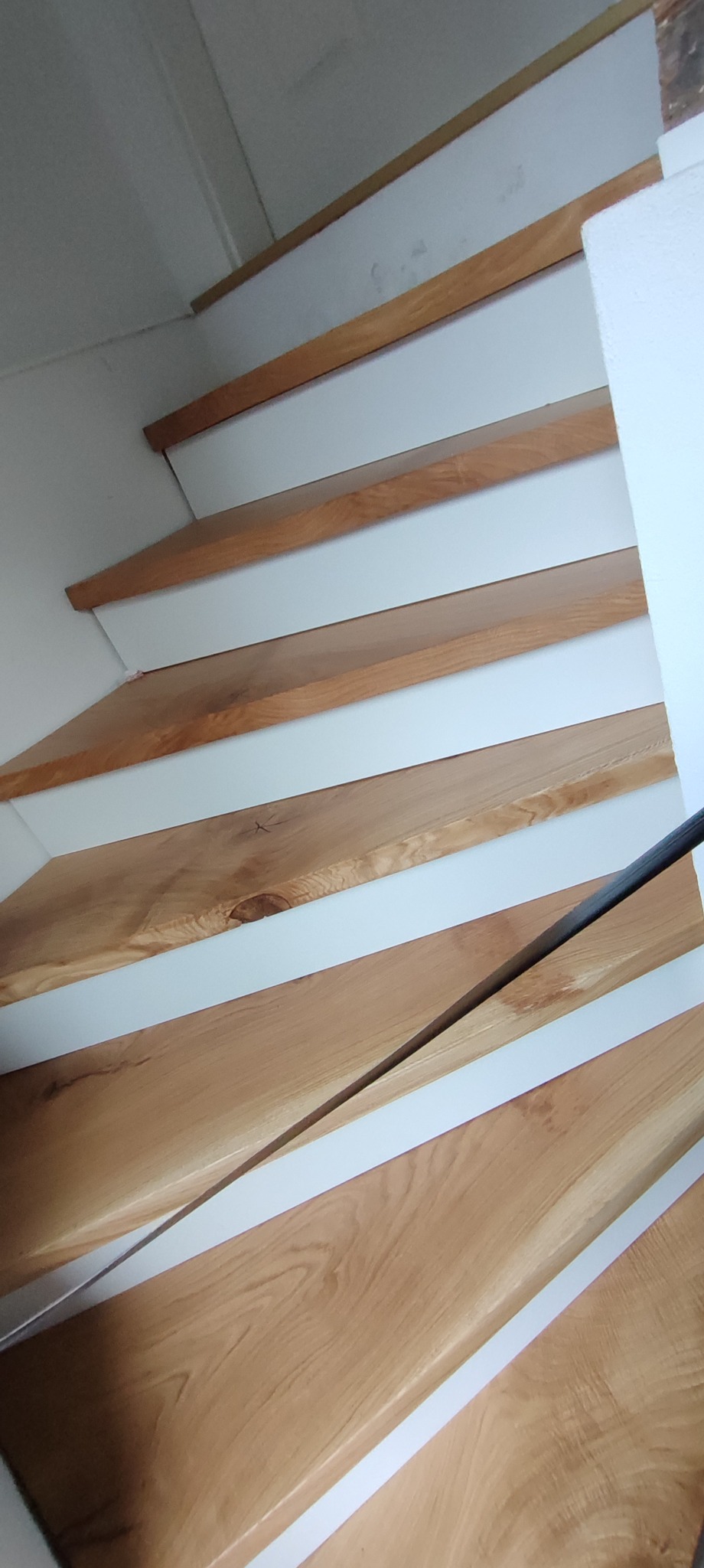 Drewniane schody z białymi podstopnicami, widok z dołu, ukazujący naturalne usłojenie drewna i minimalistyczny design. Metalowa poręcz dodaje nowoczesnego akcentu.