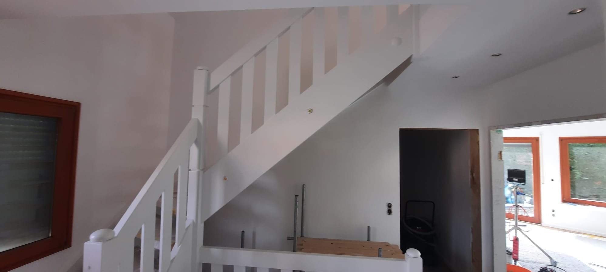 Białe schody z drewnianą balustradą w jasnym wnętrzu. Widoczne okna z brązowymi ramami i otwór drzwiowy. Ściany pomalowane na jasny kolor. Ujęcie z dołu, podkreślające wysokość schodów.