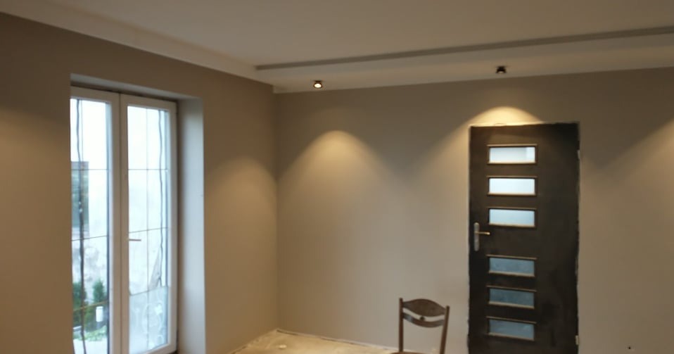 Jasne wnętrze po remoncie: świeżo pomalowane ściany w odcieniach beżu, białe listwy sufitowe, nowoczesne oświetlenie punktowe i ciemne drzwi z przeszkleniami.