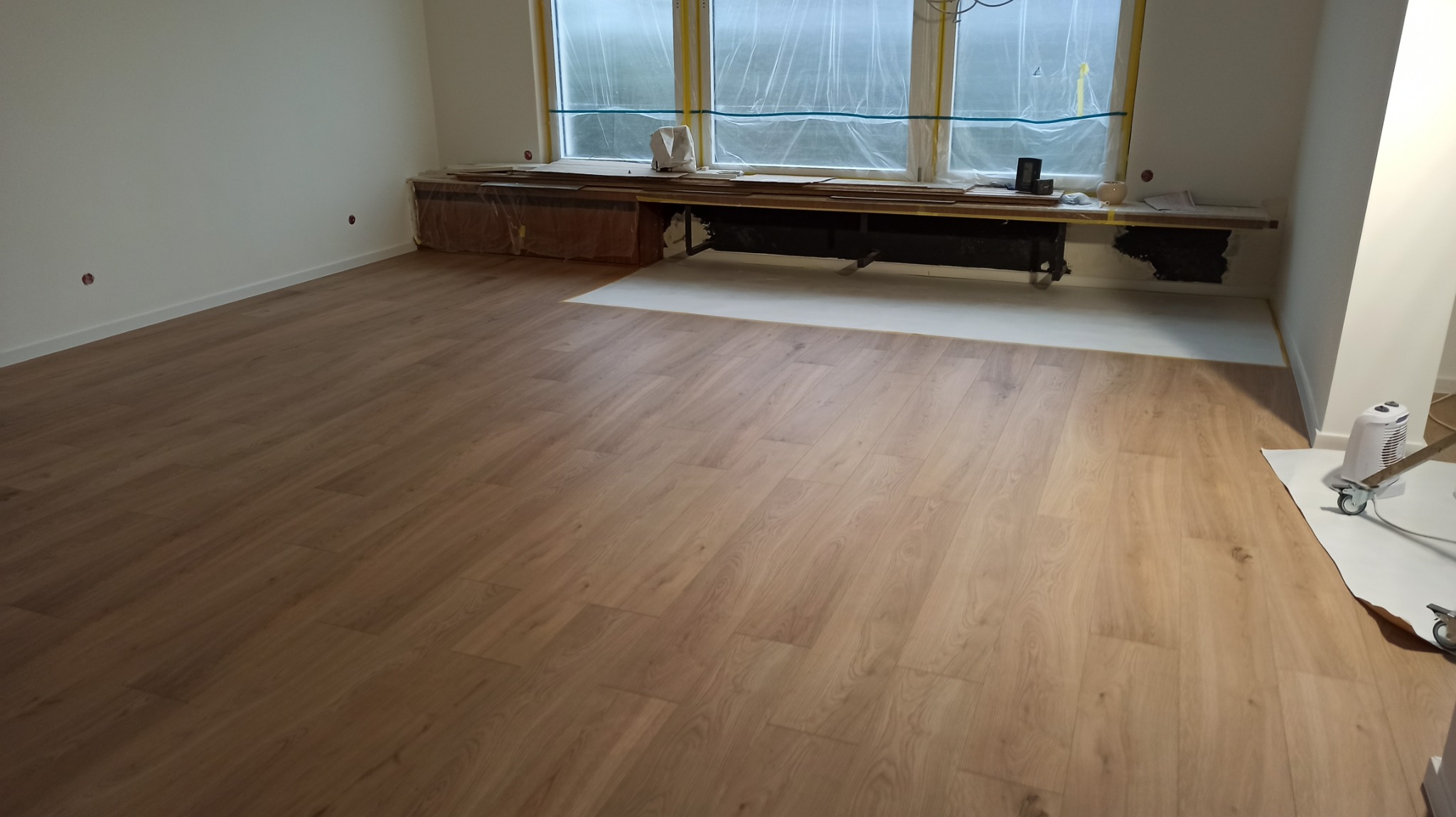 Jasne panele podłogowe ułożone w pokoju z oknem zabezpieczonym folią malarską. Fragment podłogi przy oknie jeszcze nie pokryty panelami. Widoczny grzejnik i elementy wyposażenia.