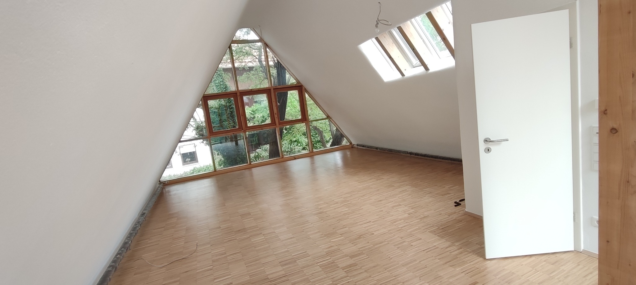 Jasne poddasze z parkietem w jodełkę, trójkątnym oknem z widokiem na zieleń i białymi ścianami. Nowe, białe drzwi po prawej stronie. Skos dachu i okno dachowe.