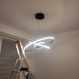 ElektroLine Marcin Fijałkowski - Nowoczesna, okrągła lampa LED zawieszona na cienkich linkach, zamontowana na suficie. Obok drabina aluminiowa, sugerująca świeży montaż. Minimalistyczny design.