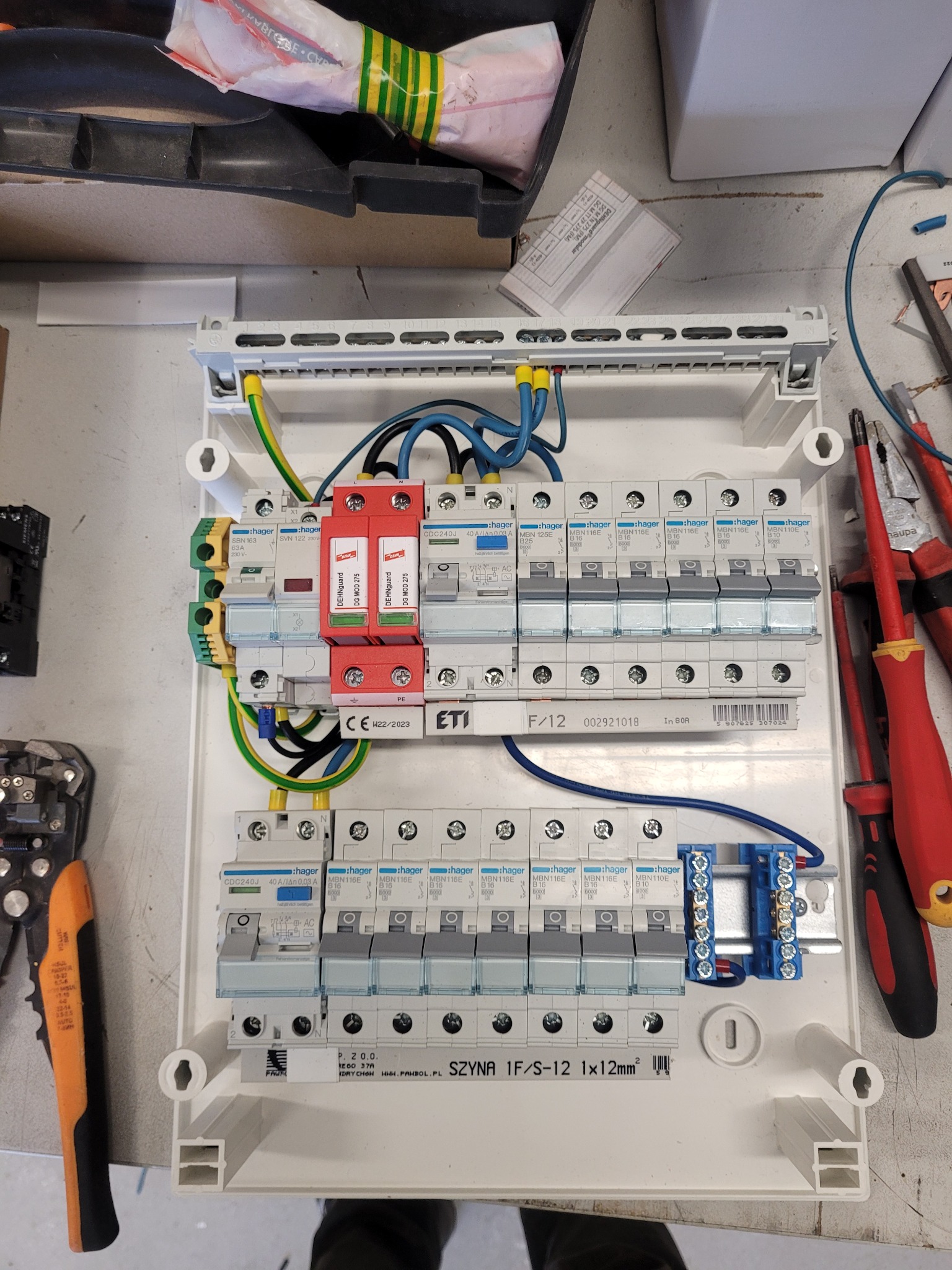 Rozdzielnica elektryczna w trakcie montażu, widoczne zabezpieczenia przepięciowe, bezpieczniki i przewody. Elementy marki Hager i ETI. Narzędzia obok.