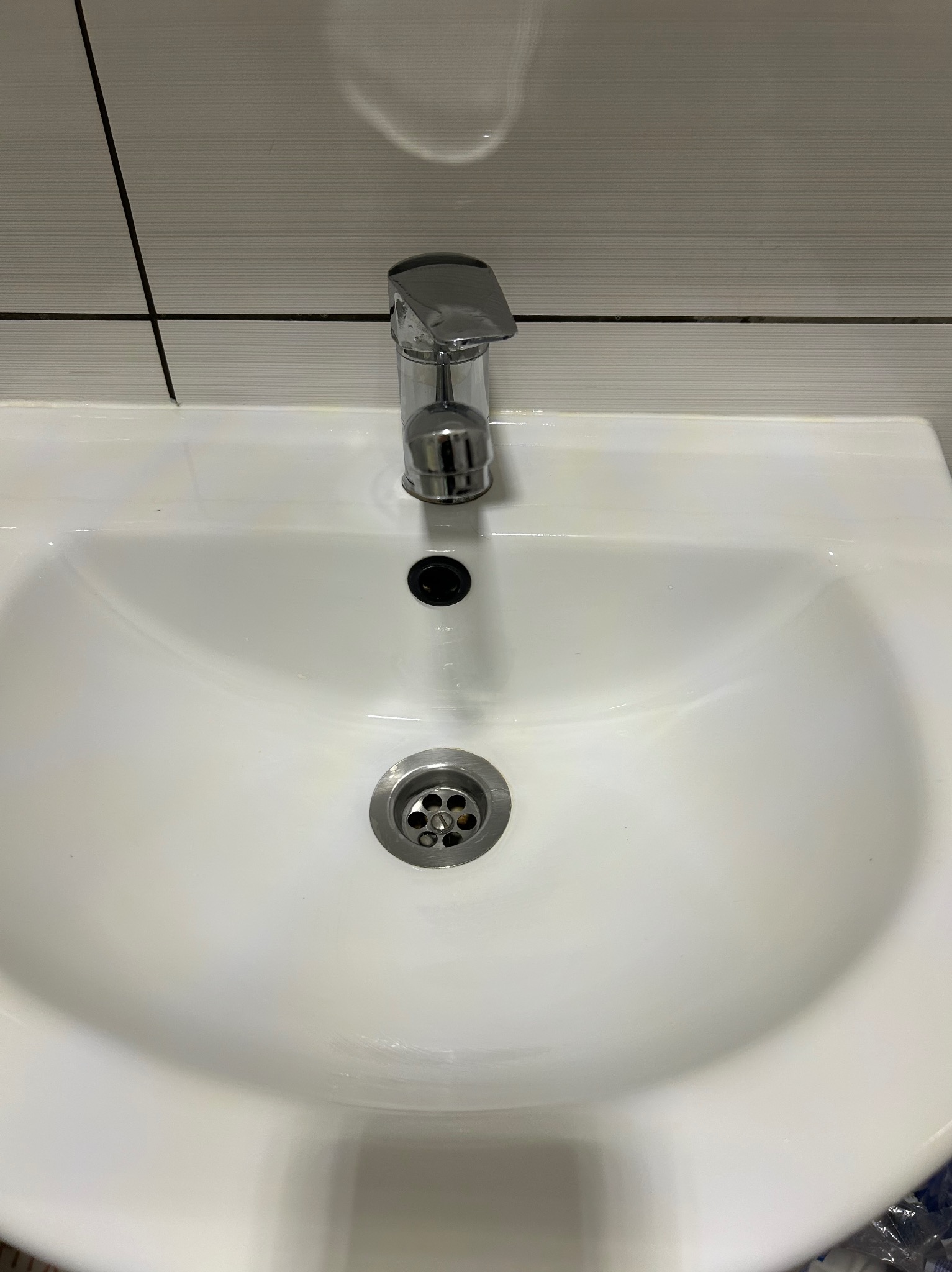 Biała umywalka z chromowaną baterią i odpływem na tle płytek. Widoczne drobne zabrudzenia, co sugeruje potrzebę wyczyszczenia.