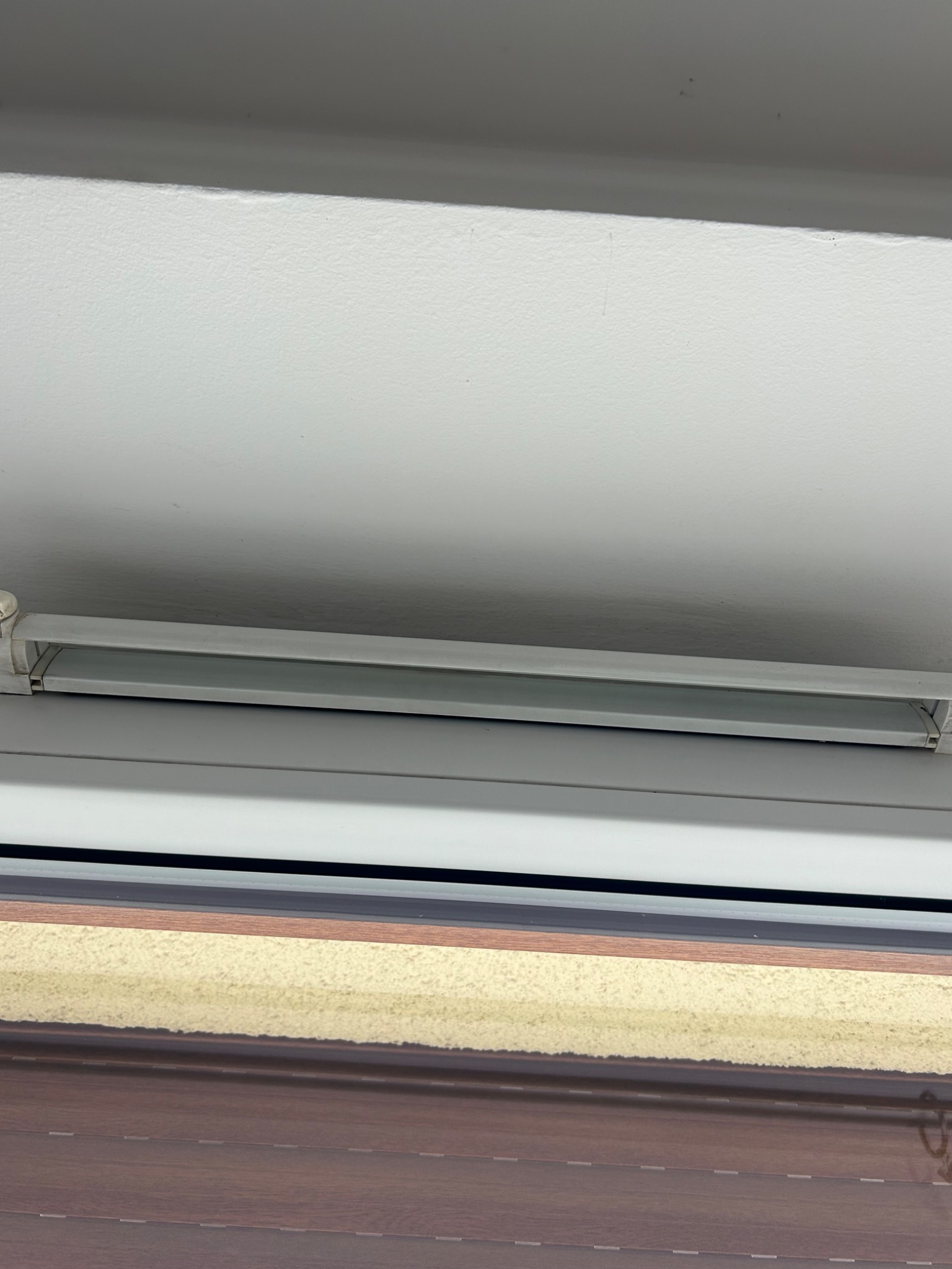 Detal okna z żaluzjami drewnopodobnymi, widoczne prowadnice i fragment ściany. Ujęcie z bliska, skupione na elementach okna.