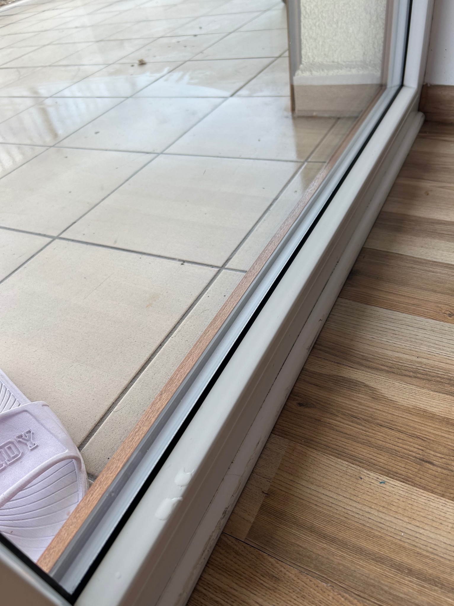 Błyszcząca, umyta podłoga z płytek i paneli, widoczna przez czystą szybę okna balkonowego. W rogu białe klapki.