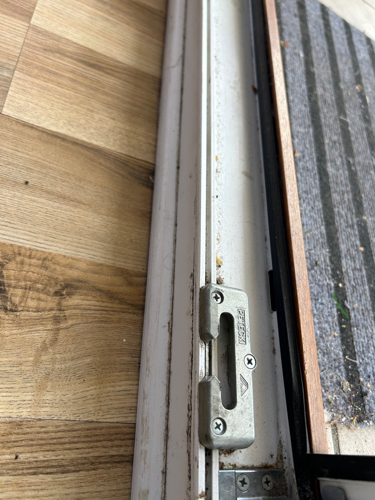 Brudna, biała rama okienna z metalowym zamkiem, widoczne zabrudzenia i kurz. Fragment podłogi z paneli i wycieraczki. Detal zaniedbanego okna.