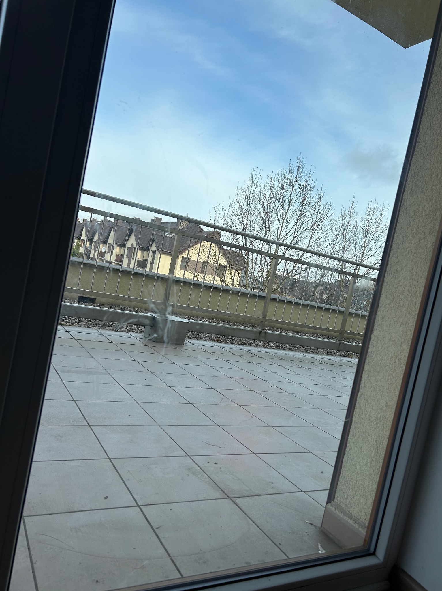 Balkon z szarymi płytkami i metalową barierką, widok z okna na budynki i drzewa. Ślady po umyciu okna widoczne na szybie.