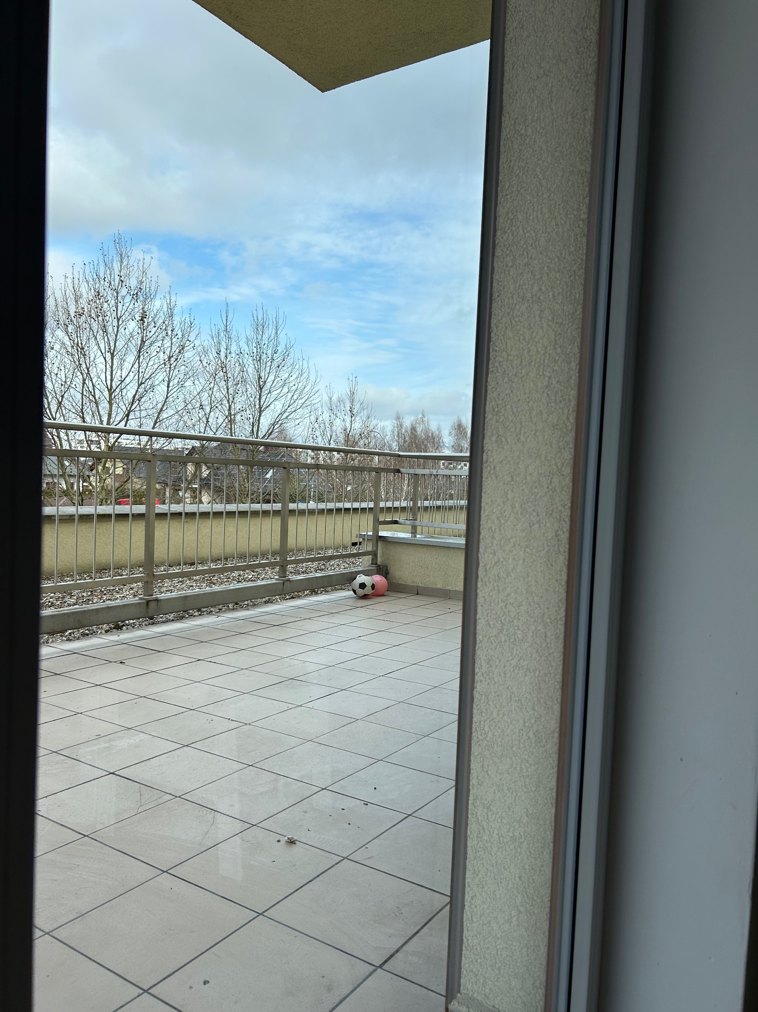 Balkon z szarymi płytkami i metalową barierką. W tle drzewa bez liści i budynki. Na podłodze leżą dwie piłki. Widok z wnętrza przez otwarte drzwi balkonowe.