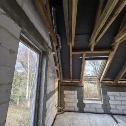 TWOJA EKIPA SPÓŁKA Z OGRANICZONĄ ODPOWIEDZIALNOŚCIĄ - Szara rura kanalizacyjna z obejmami, zamontowana pod drewnianą konstrukcją dachu na poddaszu w surowym stanie. Widoczne okna dachowe i ściany z betonu komórkowego.