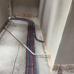 TWOJA EKIPA SPÓŁKA Z OGRANICZONĄ ODPOWIEDZIALNOŚCIĄ - Równolegle ułożone rury instalacji grzewczej w kolorach czerwonym i niebieskim, prowadzone wzdłuż ściany w surowym wnętrzu. Widoczne fragmenty podłoża i ściany w trakcie prac wykończeniowych.