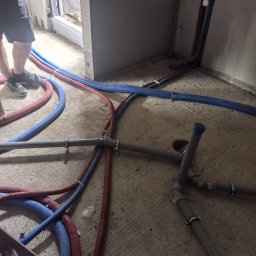 TWOJA EKIPA SPÓŁKA Z OGRANICZONĄ ODPOWIEDZIALNOŚCIĄ - Instalacja rur grzewczych i hydraulicznych w surowym wnętrzu. Rury w izolacji czerwonej i niebieskiej, szare rury PCV z łącznikami i obejmami na betonowej podłodze.