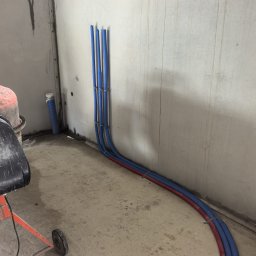 TWOJA EKIPA SPÓŁKA Z OGRANICZONĄ ODPOWIEDZIALNOŚCIĄ - Ułożone, niebiesko-czerwone rury instalacji grzewczej przy ścianie w surowym wnętrzu. Widoczna betoniarka i fragment podłogi. Etap prac przed zalaniem posadzki.