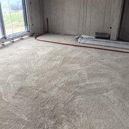 TWOJA EKIPA SPÓŁKA Z OGRANICZONĄ ODPOWIEDZIALNOŚCIĄ - Wnętrze w trakcie instalacji ogrzewania podłogowego: czerwone rurki na szarej, surowej wylewce, widoczne okno i materiały izolacyjne. Etap przed zalaniem posadzki.