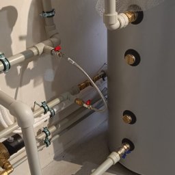 TWOJA EKIPA SPÓŁKA Z OGRANICZONĄ ODPOWIEDZIALNOŚCIĄ - Szare rury instalacji wodnej z zaworami i zbiornikiem, zamontowane przy ścianie. Widoczne złączki, mocowania i fragment podłogi. Detal instalacji.