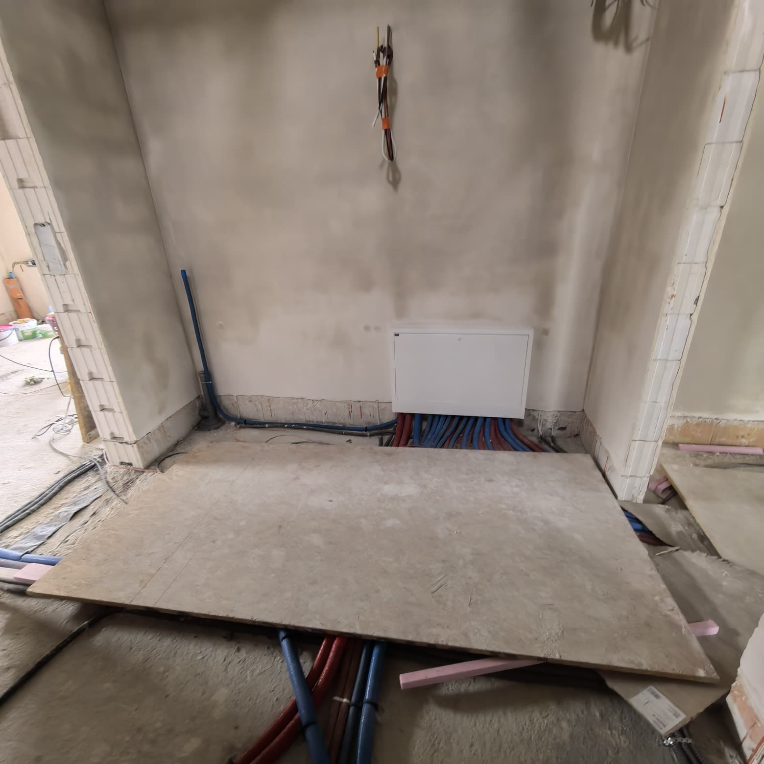 Instalacja wodna i ogrzewania podłogowego w surowym wnętrzu. Widoczne rury w kolorach niebieskim i czerwonym, prowadzone pod płytą OSB. Biała skrzynka na ścianie.