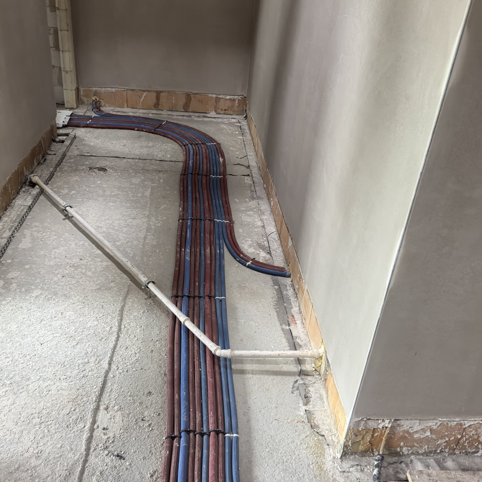 Równolegle ułożone rury instalacji grzewczej w kolorach czerwonym i niebieskim, prowadzone wzdłuż ściany w surowym wnętrzu. Widoczne fragmenty podłoża i ściany w trakcie prac wykończeniowych.
