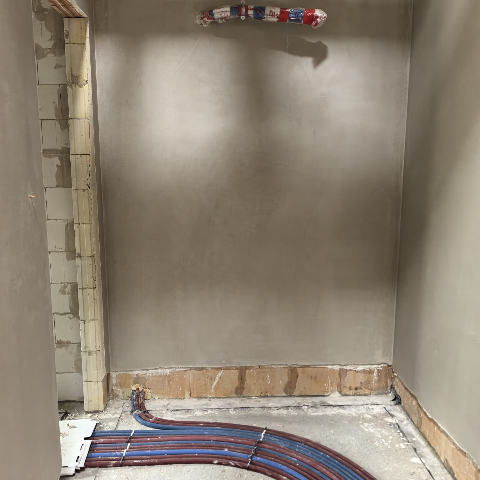 Wnętrze w trakcie prac hydraulicznych: rury ułożone przy podłodze i wyprowadzone na ścianie, ściany otynkowane, widoczne bloczki. Etap instalacji przed wykończeniem.