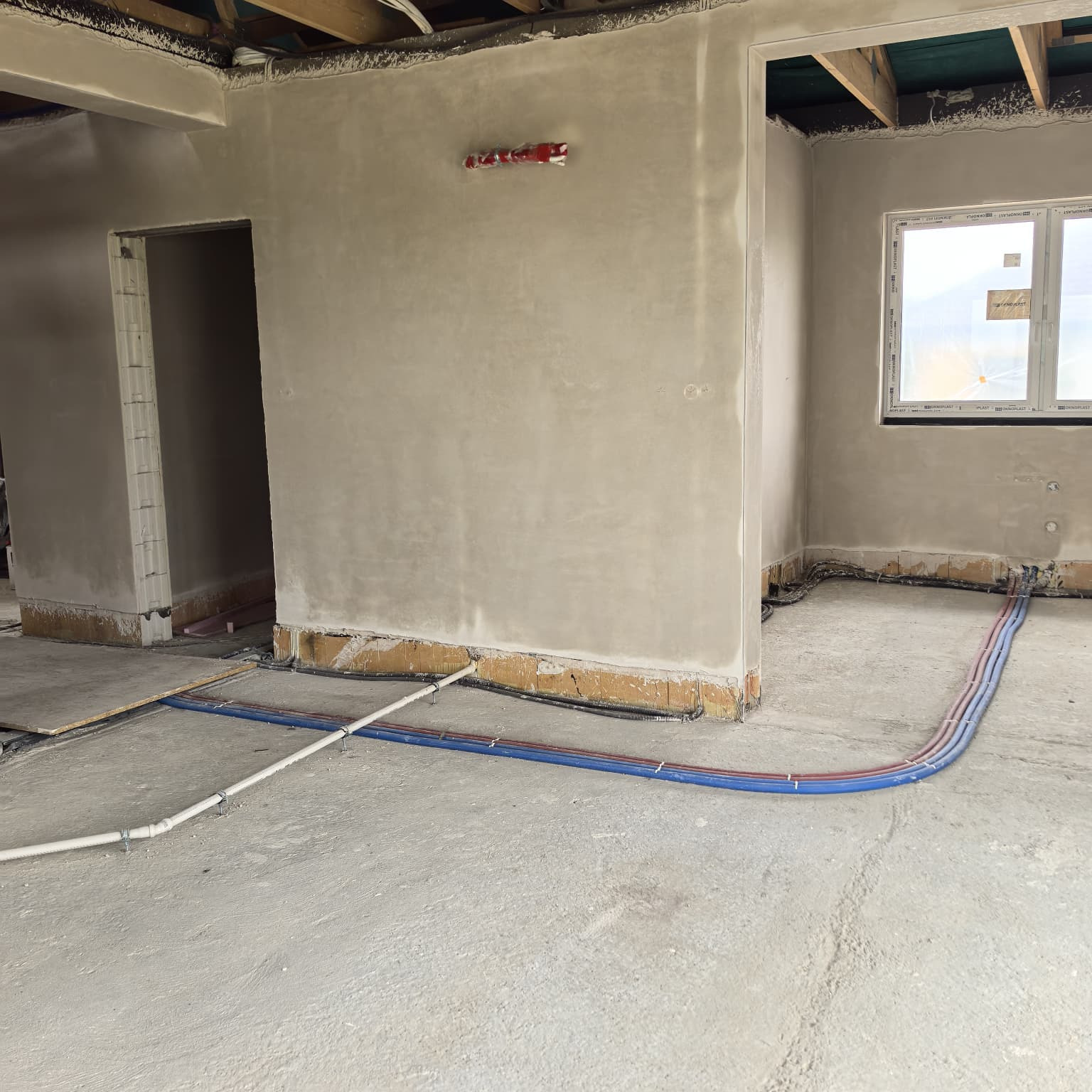 Instalacja rur grzewczych i wodnych w surowym stanie budynku we Wrocławiu. Widoczne rury w kolorze niebieskim, czerwonym i białym, prowadzone po podłodze.