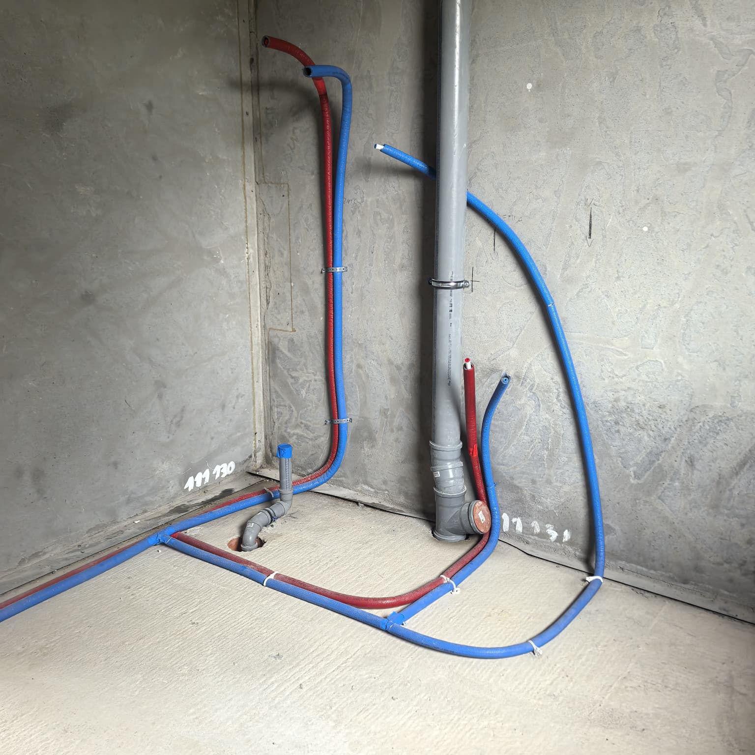 Instalacja hydrauliczna w surowym wnętrzu: rury PEX (czerwone i niebieskie) prowadzone wzdłuż ścian i podłogi, szara rura kanalizacyjna pionowo. Etap prac instalacyjnych.