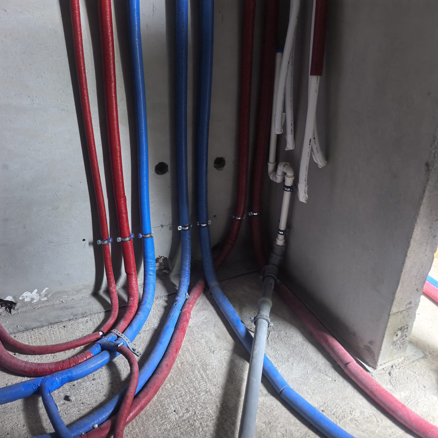 Instalacja wodna w surowym wnętrzu: czerwone i niebieskie rury prowadzone wzdłuż szarej ściany, z widocznymi mocowaniami i fragmentami białych rur. Surowy beton w tle.