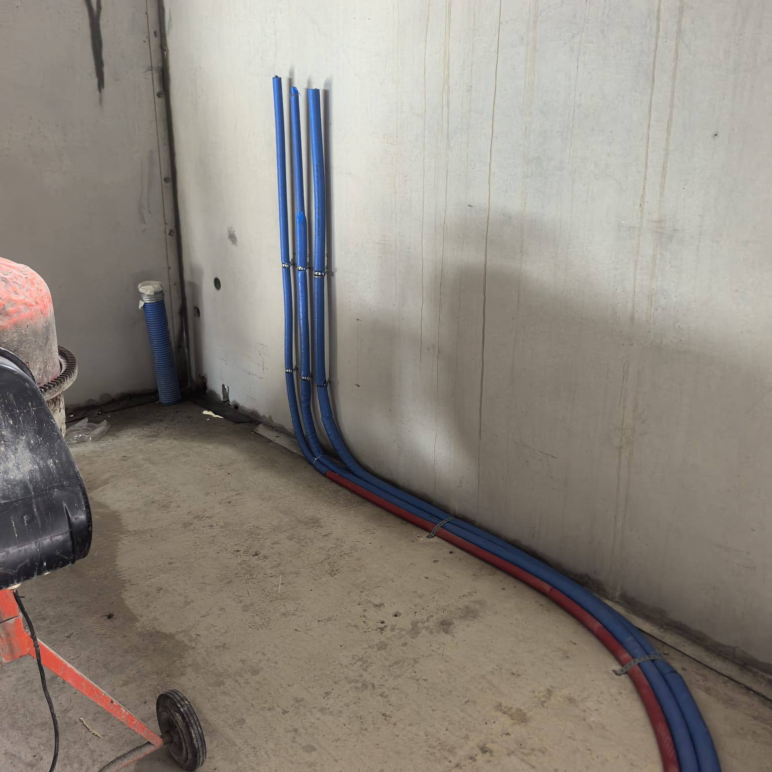 Ułożone, niebiesko-czerwone rury instalacji grzewczej przy ścianie w surowym wnętrzu. Widoczna betoniarka i fragment podłogi. Etap prac przed zalaniem posadzki.