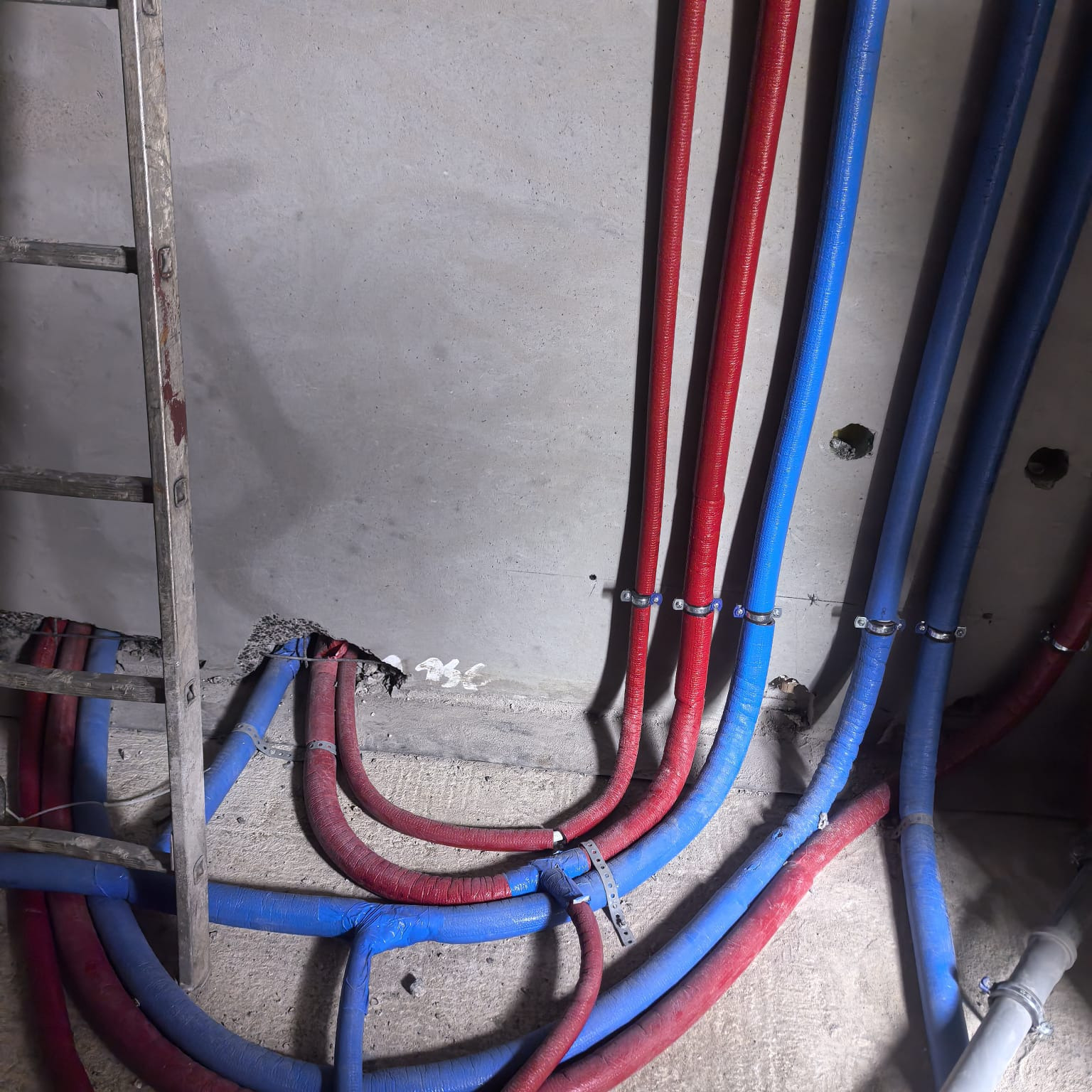 Czerwone i niebieskie rury instalacji grzewczej/hydraulicznej, przymocowane do surowej ściany, widoczne fragmenty drabiny. Instalacja w trakcie montażu.