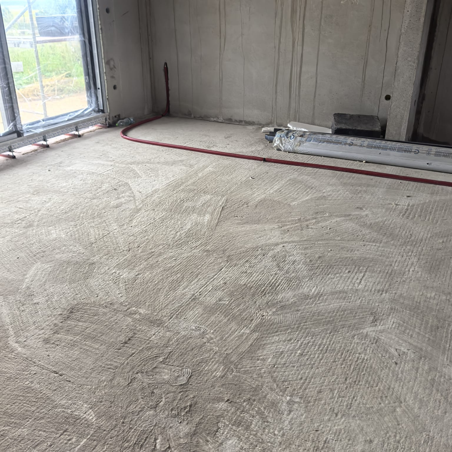 Wnętrze w trakcie instalacji ogrzewania podłogowego: czerwone rurki na szarej, surowej wylewce, widoczne okno i materiały izolacyjne. Etap przed zalaniem posadzki.