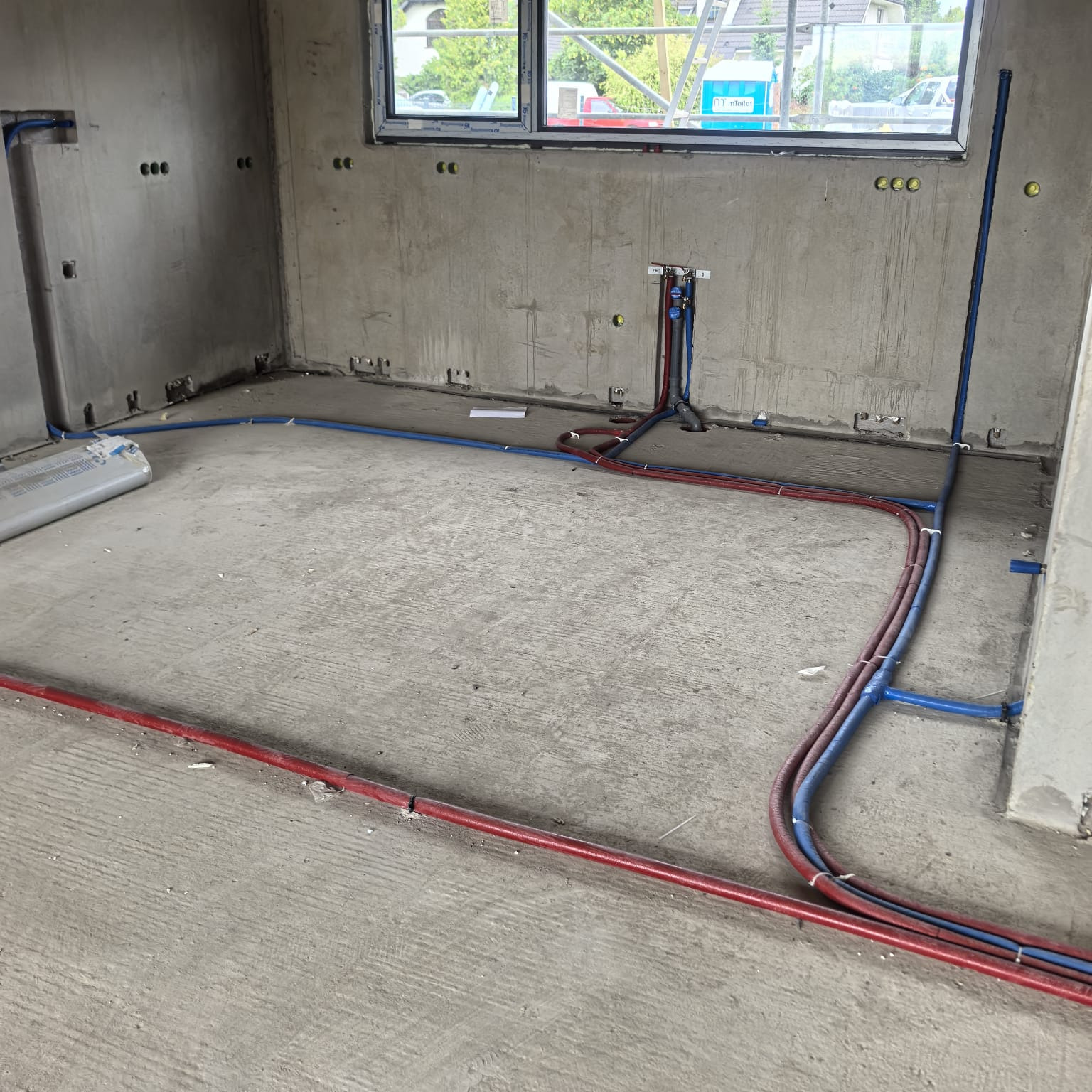 Surowy stan: czerwone i niebieskie rury instalacji grzewczej/hydraulicznej ułożone na szarej, betonowej podłodze w pomieszczeniu z oknem i surowymi ścianami.