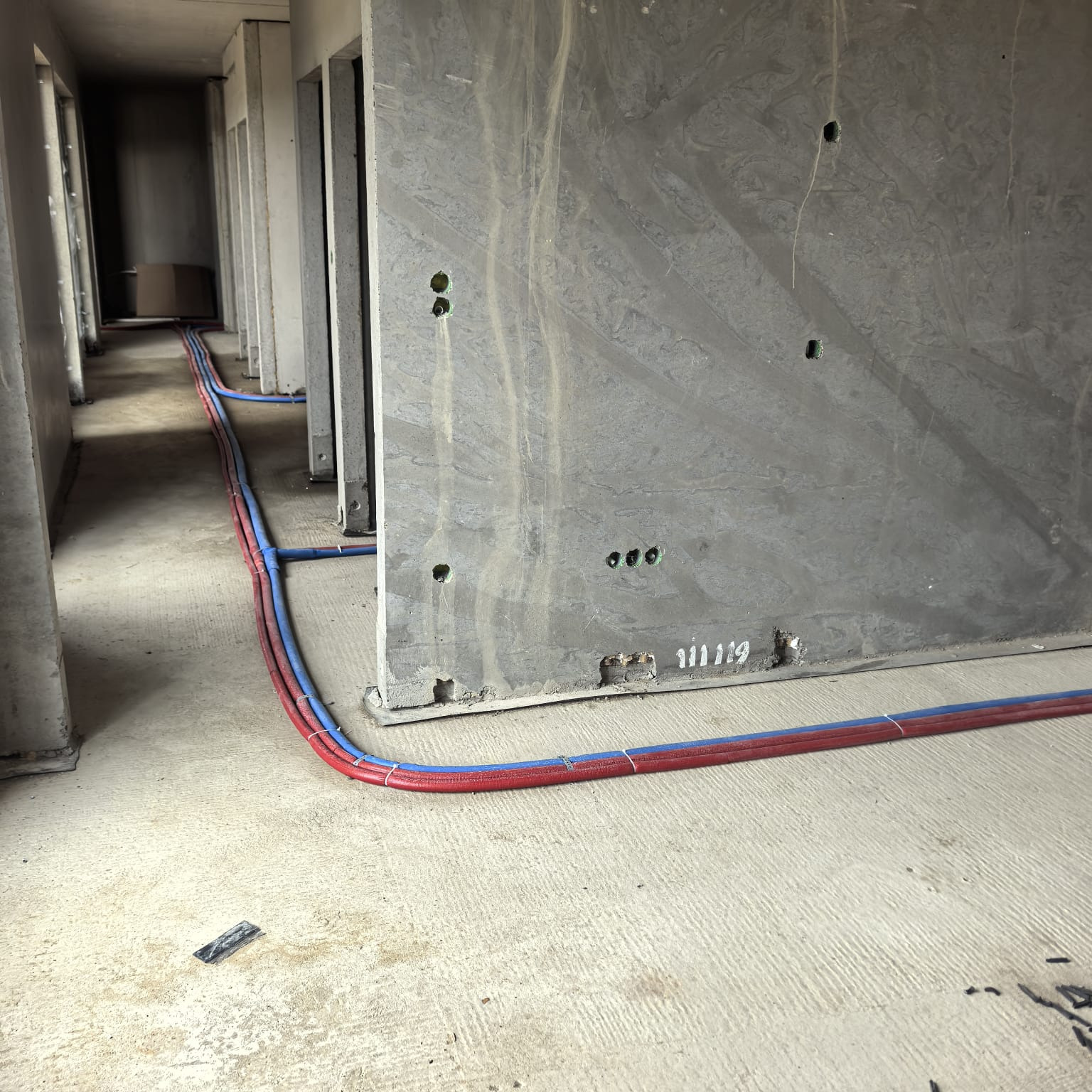 Instalacja rur grzewczych (czerwone i niebieskie) w surowym wnętrzu budynku w trakcie budowy. Widoczne betonowe ściany i podłoga. Rury ułożone wzdłuż ściany.