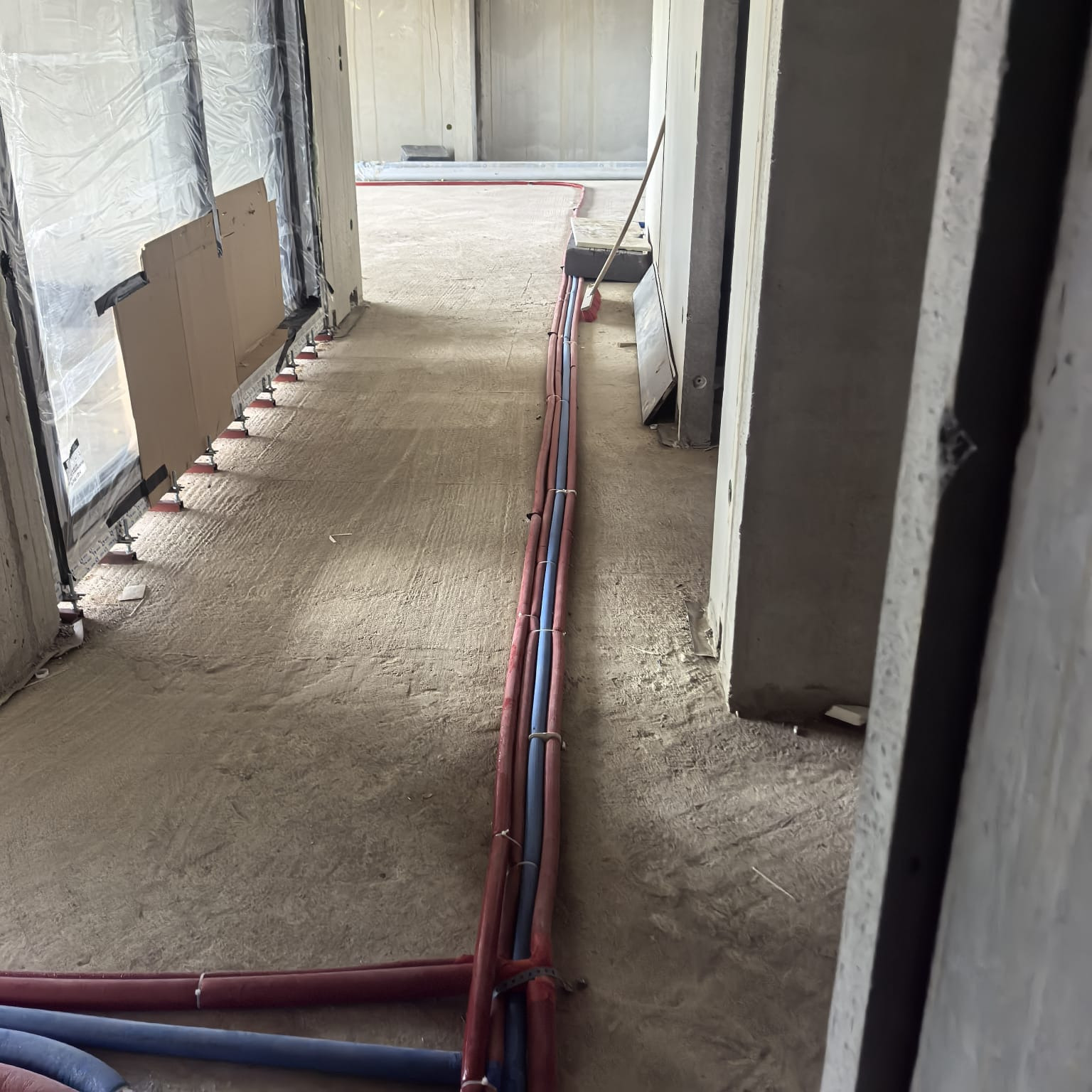 Instalacja rur (czerwone i niebieskie) na podłodze w surowym stanie, widok z perspektywy korytarza. Rury prowadzone wzdłuż ściany, przygotowane do dalszych prac wykończeniowych.