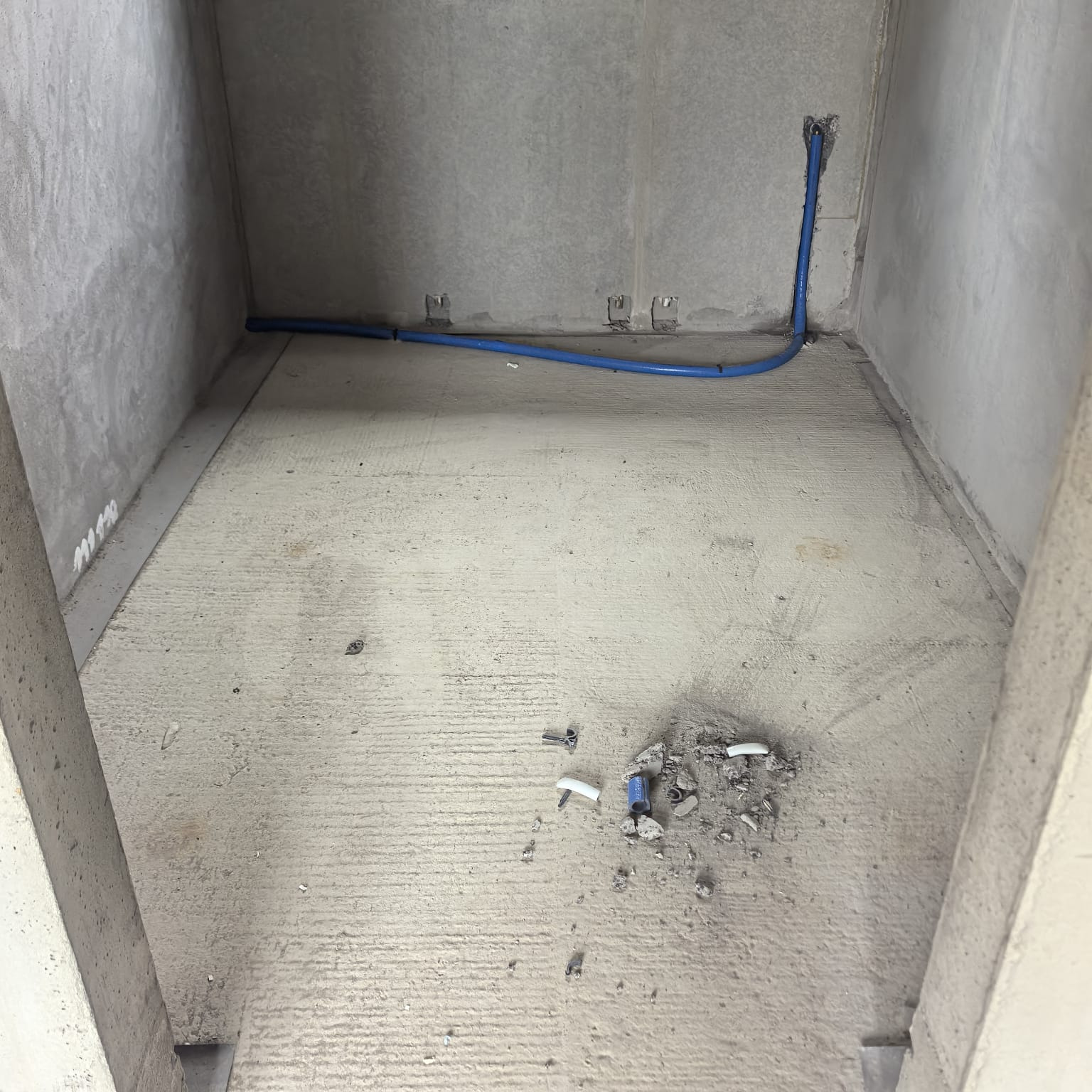 Surowy betonowy pokój z niebieskimi rurami instalacji wodnej ułożonymi wzdłuż ścian i podłogi. Widoczne ślady po kuciu i montażu.