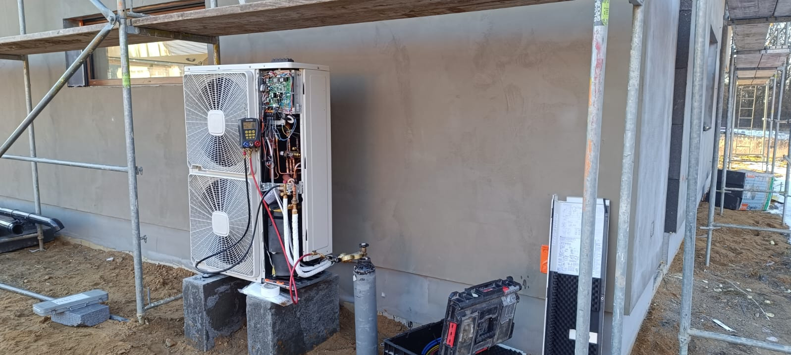 Biała pompa ciepła typu split na zewnątrz budynku w trakcie montażu. Widoczne narzędzia i otwarte panele urządzenia. Budynek w stanie surowym, rusztowania.