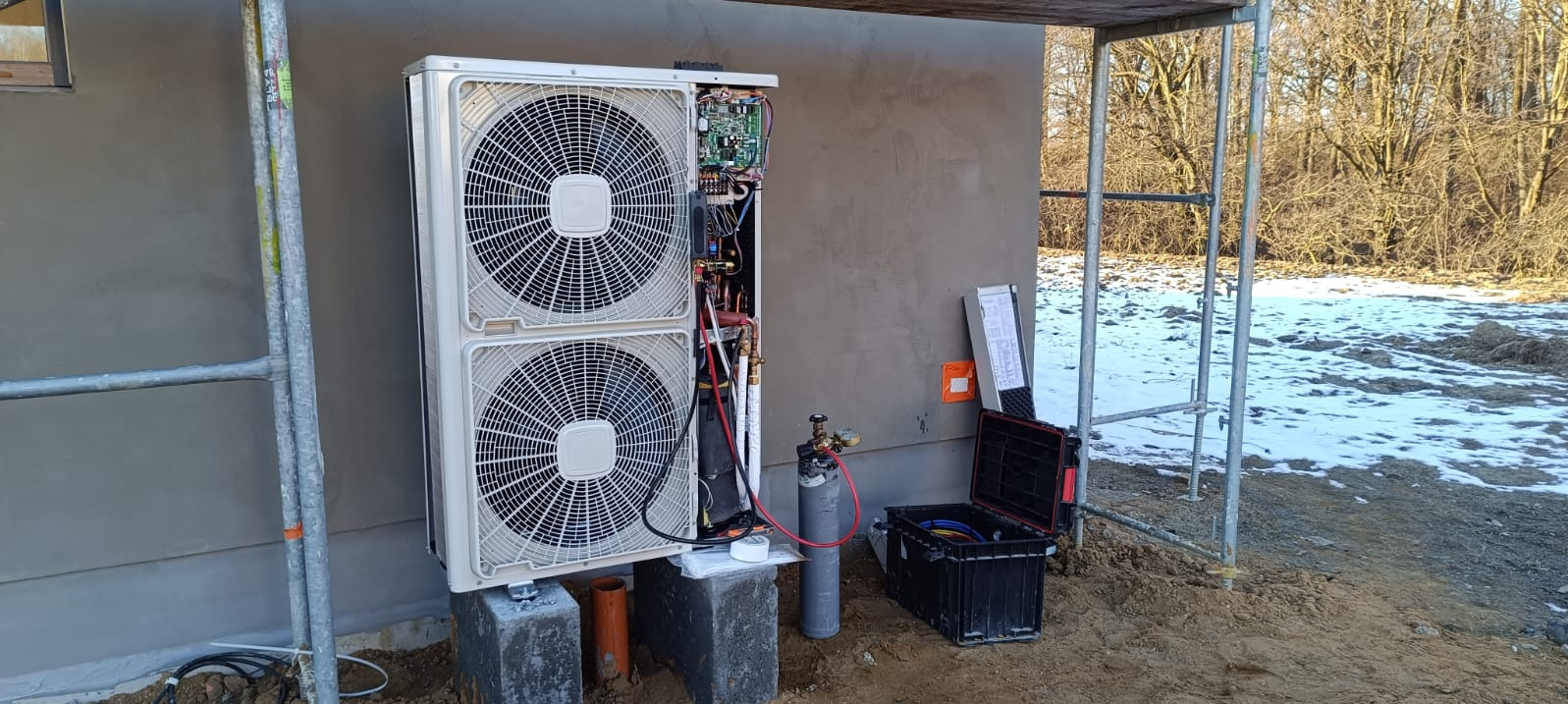 Biała pompa ciepła typu split, zamontowana na zewnątrz budynku w trakcie instalacji. Widoczne narzędzia, butla z gazem i skrzynka z przewodami. Śnieg na ziemi.