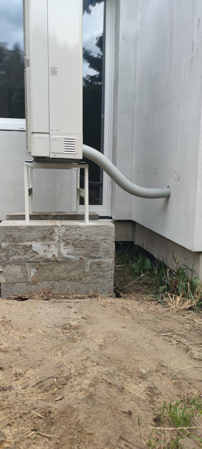 Zewnętrzna jednostka klimatyzacji zamontowana na betonowym fundamencie, z widocznym przewodem odprowadzającym skropliny przez ścianę budynku. Widok z dołu.