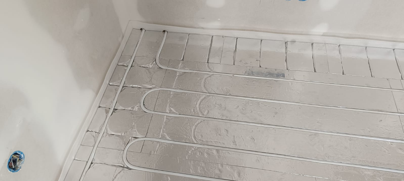 Ułożona instalacja ogrzewania podłogowego na folii aluminiowej, widok z góry, rury w kształcie pętli, przygotowane do zalania posadzką. W tle ściana w surowym stanie.