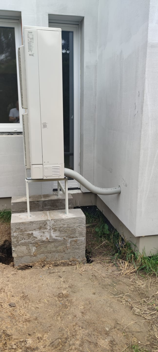 Zewnętrzna jednostka klimatyzacji zamontowana na betonowym fundamencie przy ścianie budynku. Szara rura łączy jednostkę ze ścianą. Widoczne okno w tle.