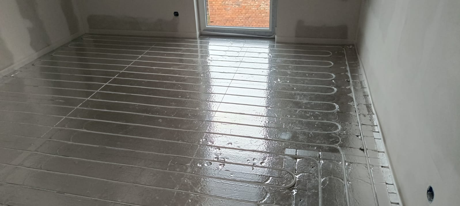 Błyszcząca folia aluminiowa z rurami ogrzewania podłogowego w surowym wnętrzu. Widok na okno z ceglanym budynkiem w tle. Etap instalacji przed wylewką.