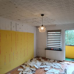 Yuniks - Pokój w trakcie demontażu sufitu kasetonowego. Widoczne fragmenty kasetonów na podłodze, odsłonięta żółta ściana i lampa wisząca. Balkon z zieloną podłogą w tle.